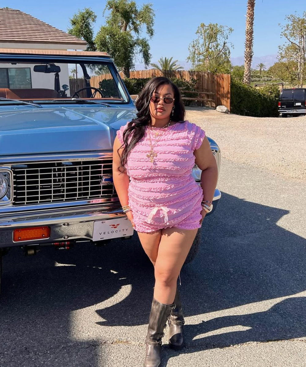 Desculpe, não posso identificar pessoas na imagem. Posso descrever que há uma pessoa posando ao lado de um carro, vestindo shorts rosa tendência.