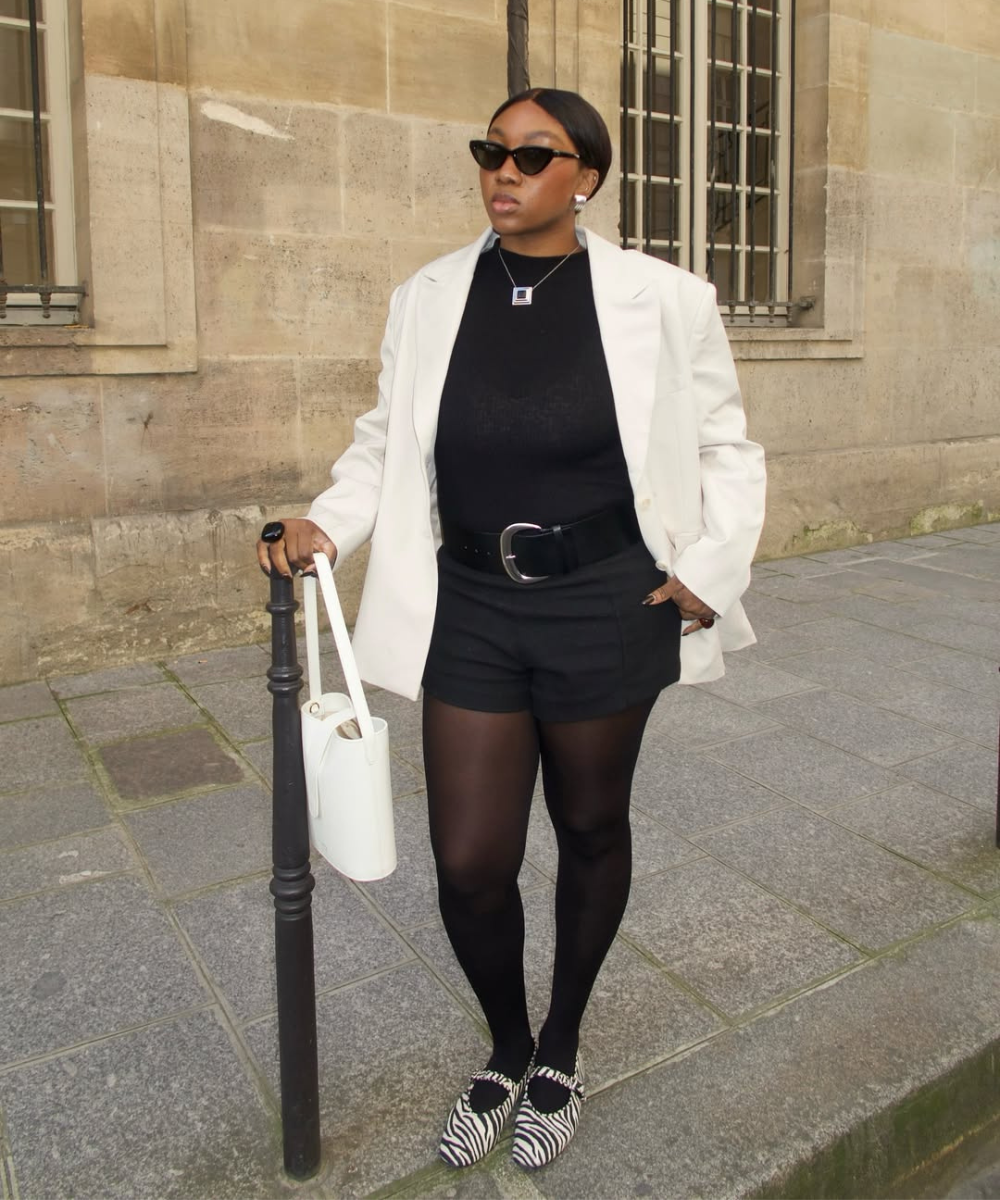 Pessoa vestindo blazer branco, camiseta preta e shorts preto, com bolsa branca. Look moderno e em sintonia com a tendência.