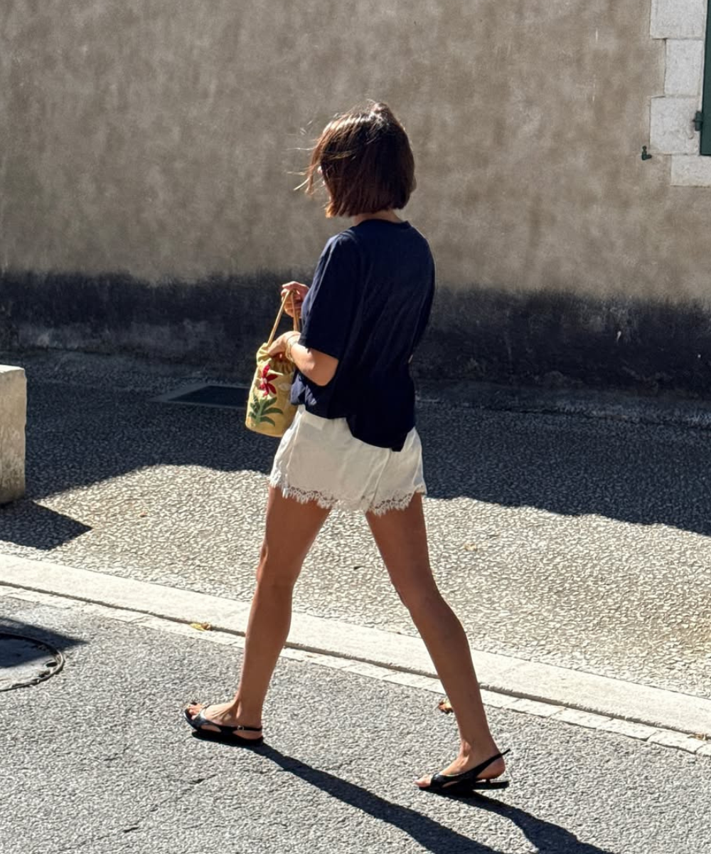 Pessoa caminha com short branco de renda, blusa escura e bolsa colorida. Shorts tendência em evidência.
