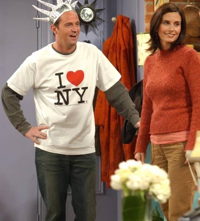 Homem com uma camisa i love new york com coração no meio. O homem veste um chapéu de papel ao lado de uma mulher sorrindo em ambiente interno.