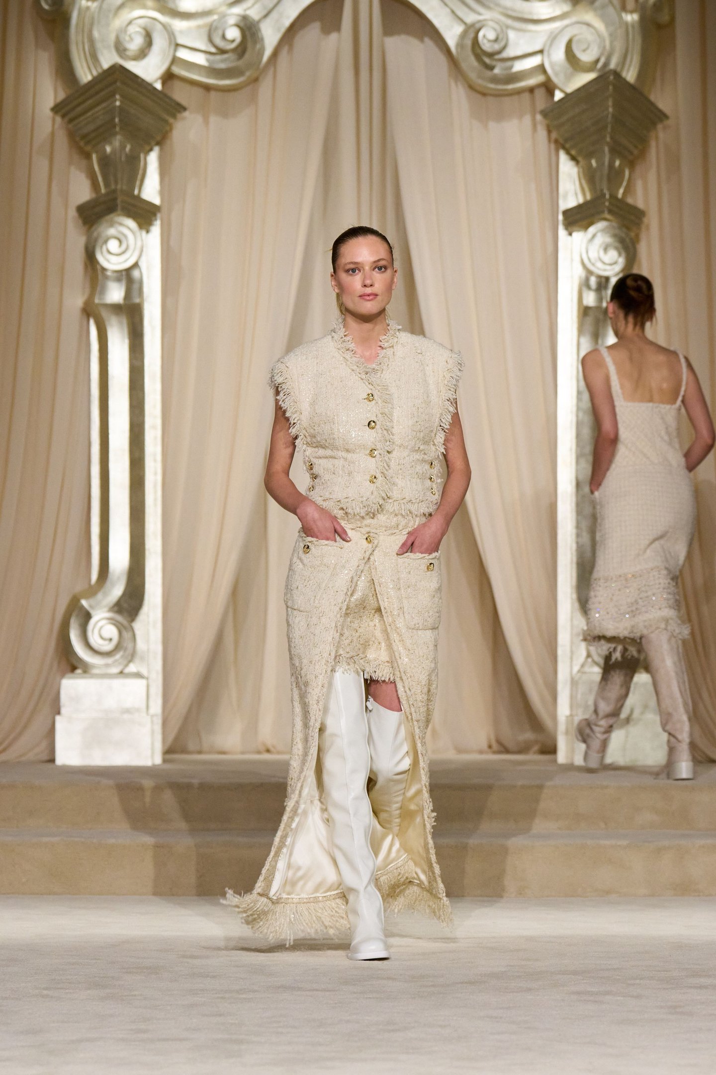 Modelo usando conjunto bege com detalhes em franjas e botas brancas em desfile de alta-costura. Cortinas luxuosas compõem o fundo.