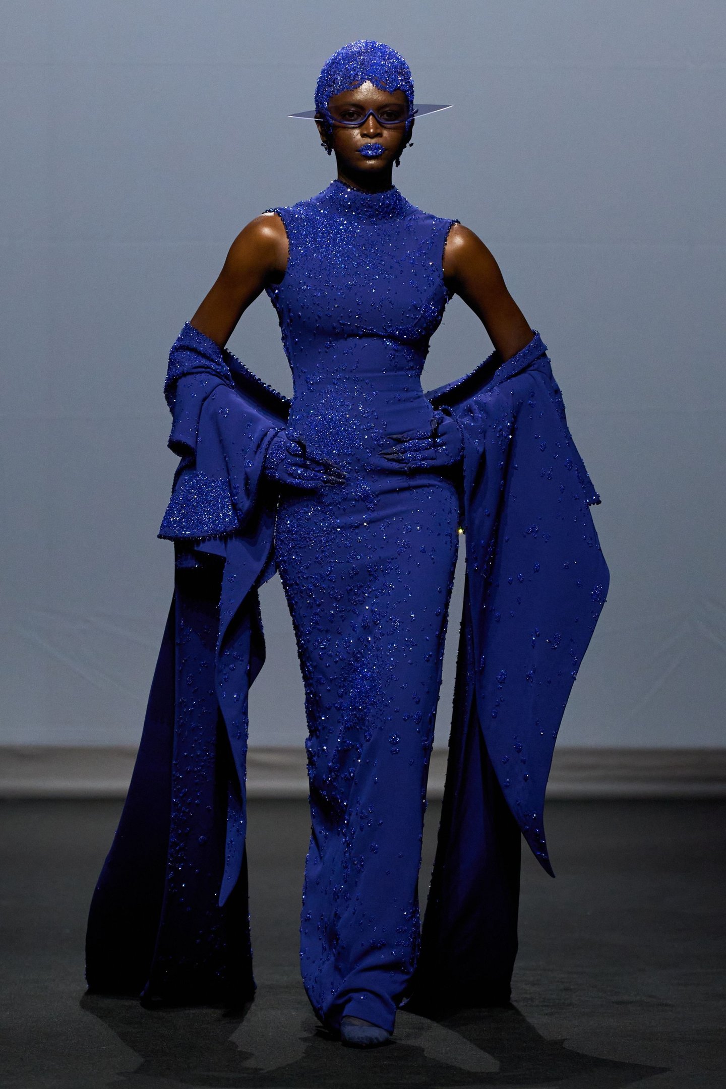 Modelo em vestido azul vibrante com cristais, usando adereços futuristas. Estilo reflete tendências da semana de alta-costura.
