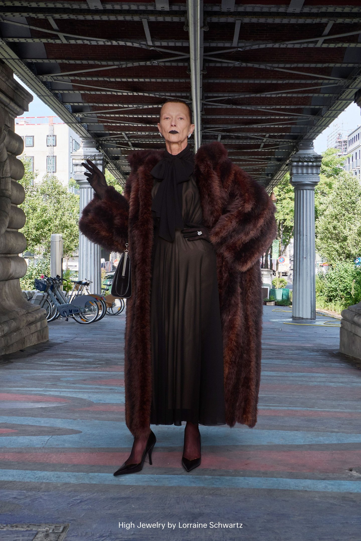 Uma pessoa em capa de pele marrom e vestido preto, tendência da alta-costura, embaixo de uma ponte.