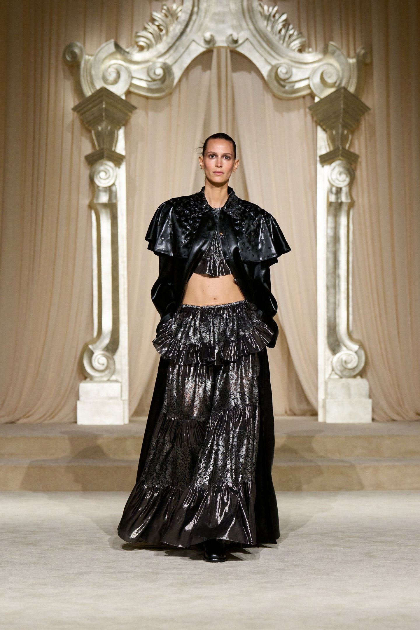 Modelo em passarela vestindo traje sofisticado preto com capa e saia volumosa, em desfile de alta-costura.