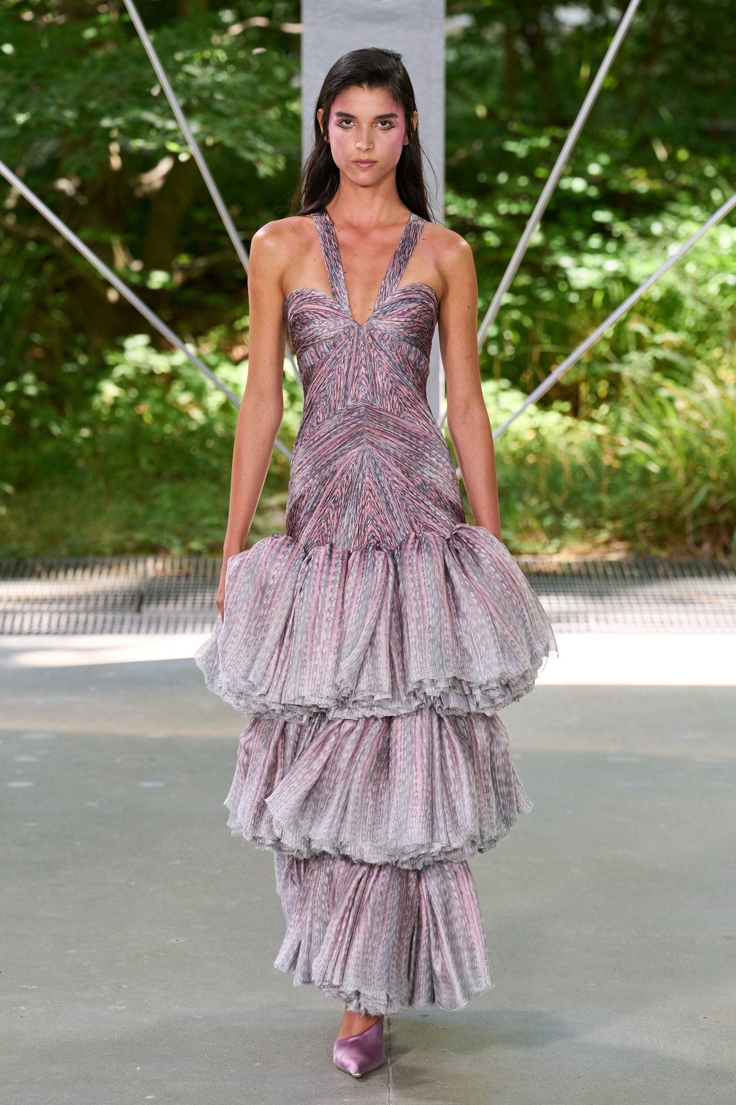 Modelo usando vestido longo texturizado com babados em tons de rosa e lilás desfila em meio a um ambiente natural. Alinhado às tendências da semana de alta-costura.