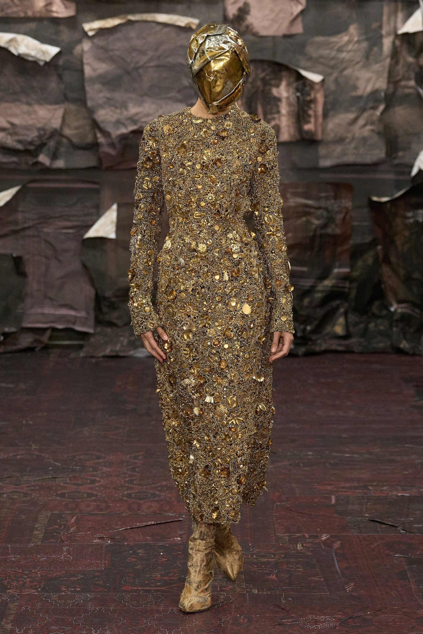 Vestido adornado com bordados dourados e máscara metalizada em passarela, influências das tendências de alta-costura.