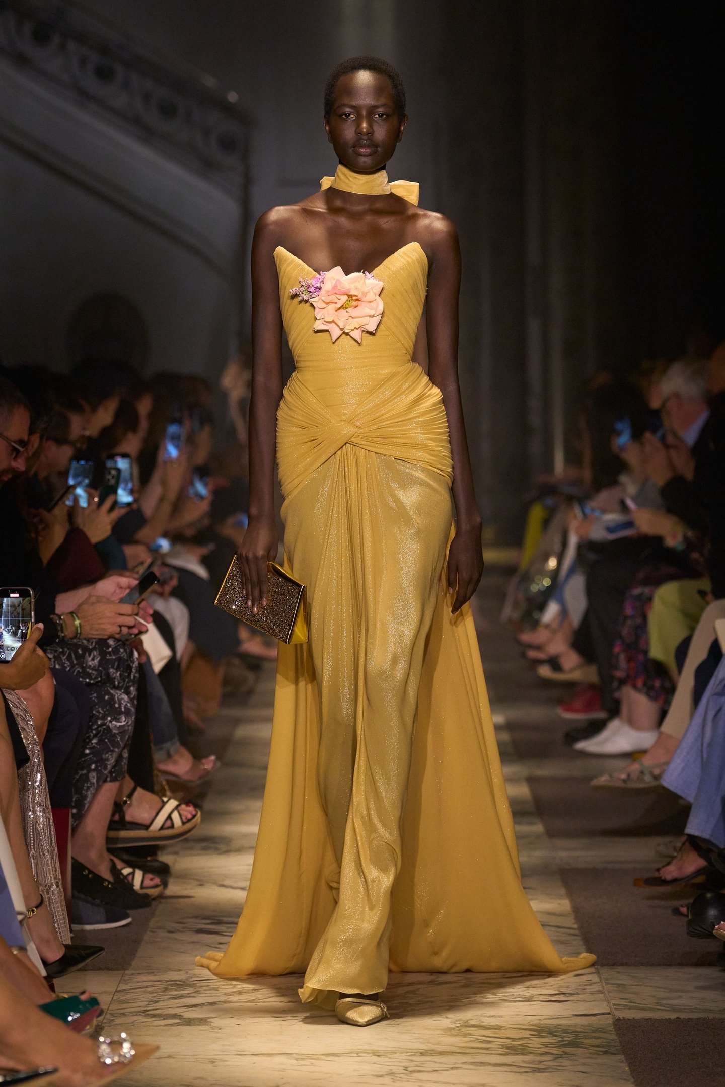 Modelo exibe vestido amarelo drapeado com flor aplicada, refletindo tendências da semana de alta-costura.