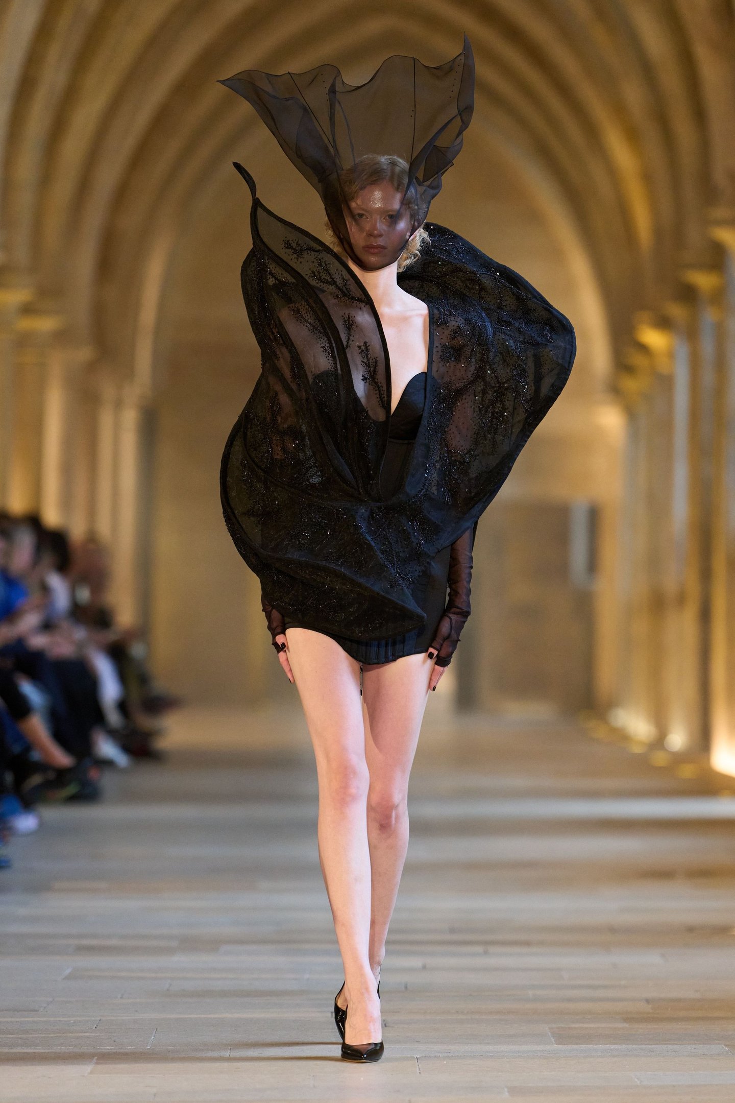 Modelo em desfile usa vestido preto com design avant-garde, refletindo tendências atuais da semana de alta-costura.