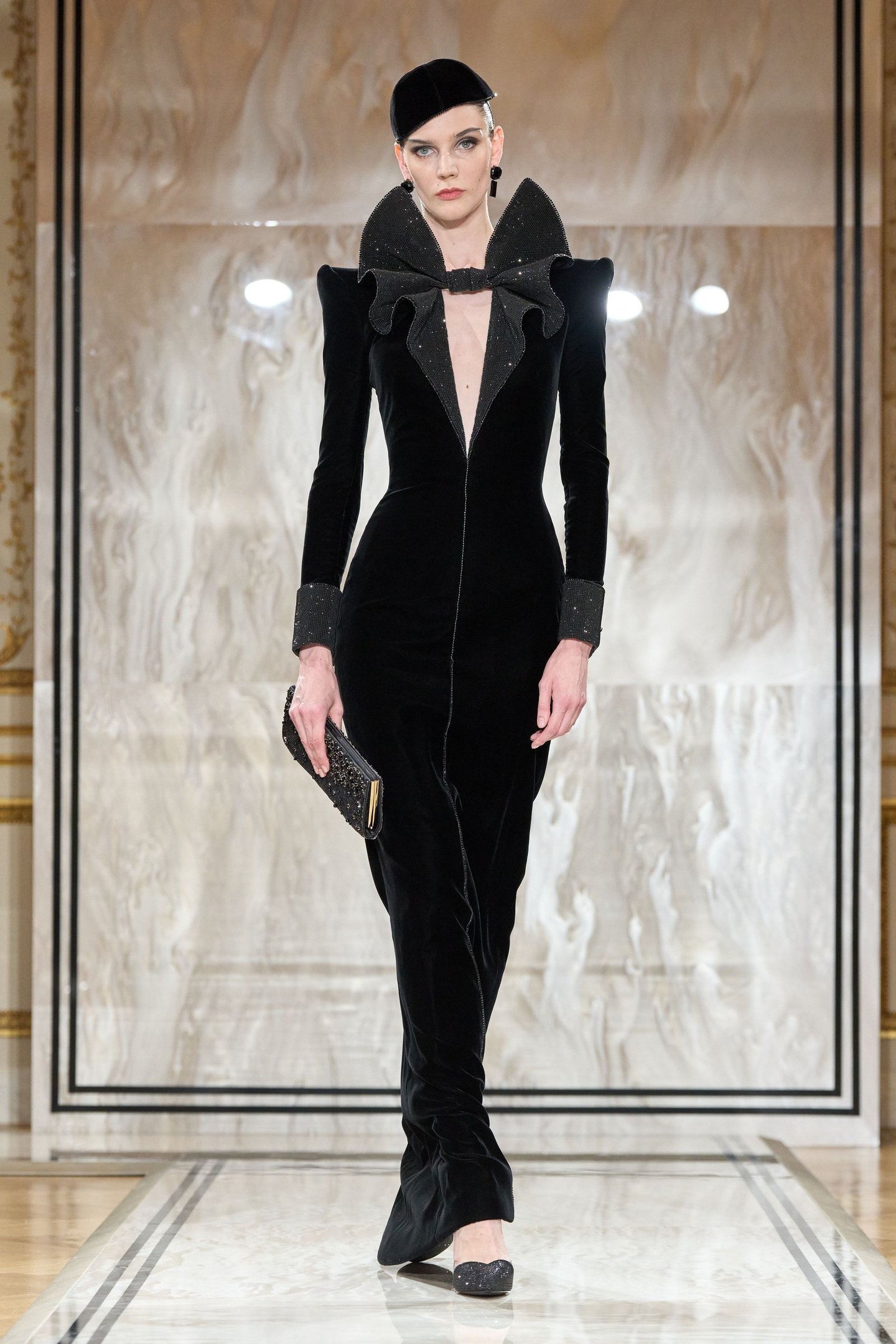 Modelo veste elegante vestido preto com gola alta volumosa e detalhes brilhantes, refletindo tendências da semana de alta-costura.