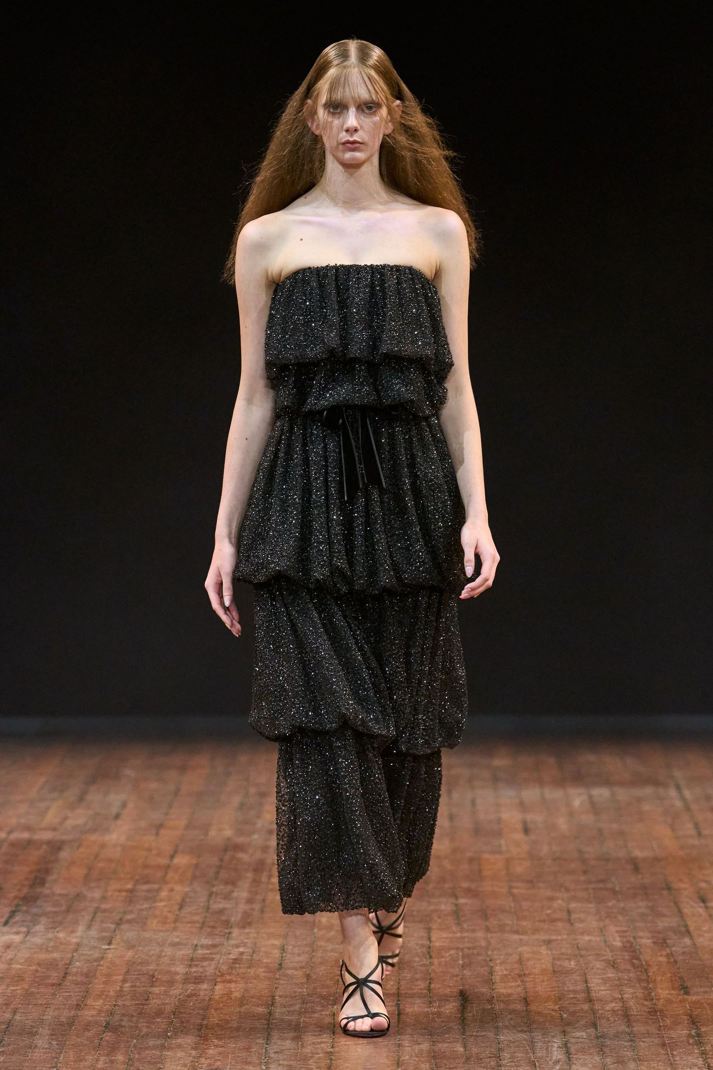 Modelo em desfile usando vestido preto brilhante com babados, destacando tendências da semana de alta-costura.