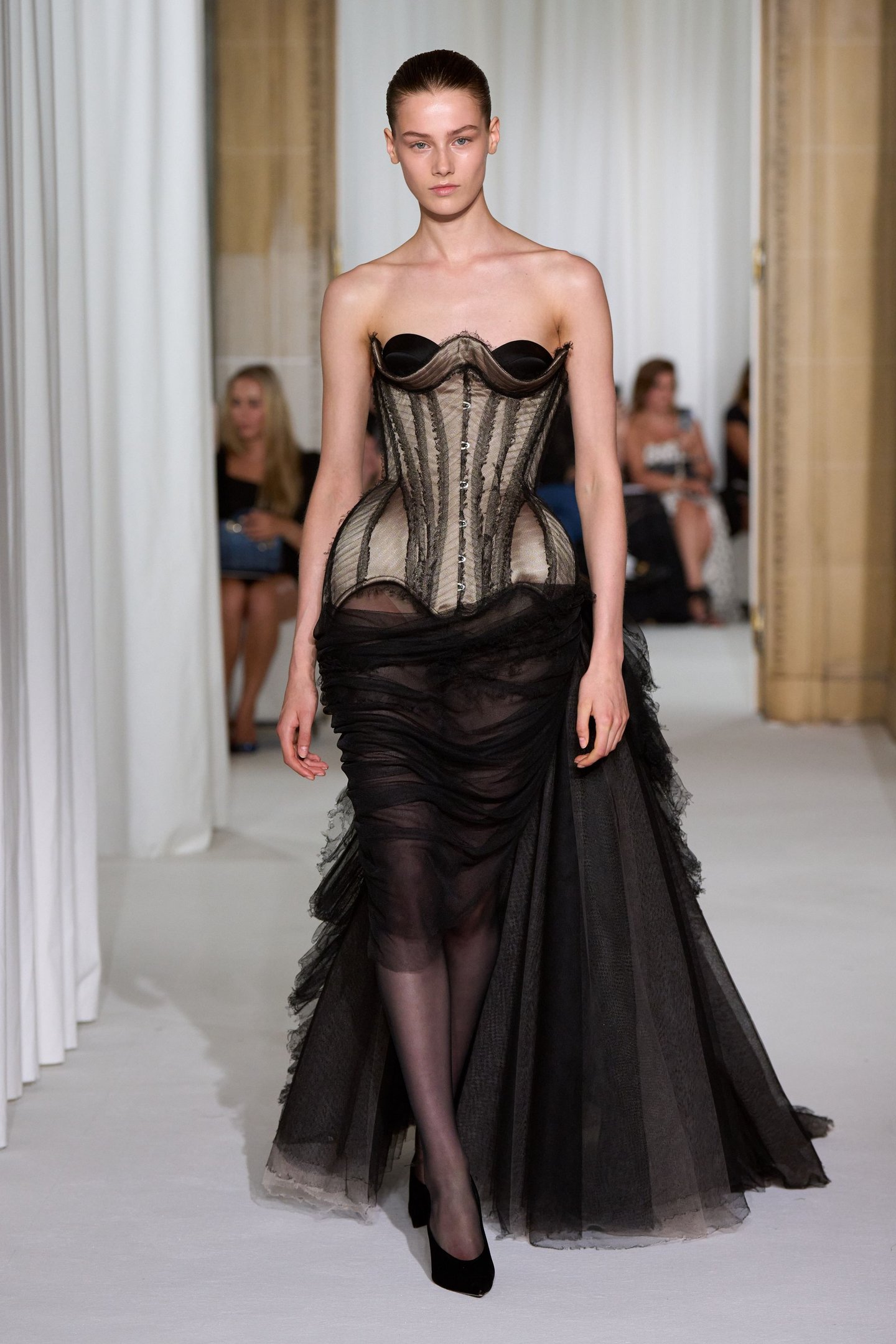 Modelo usa vestido preto com corpete estruturado e saia transparente, refletindo tendências da semana de alta-costura.
