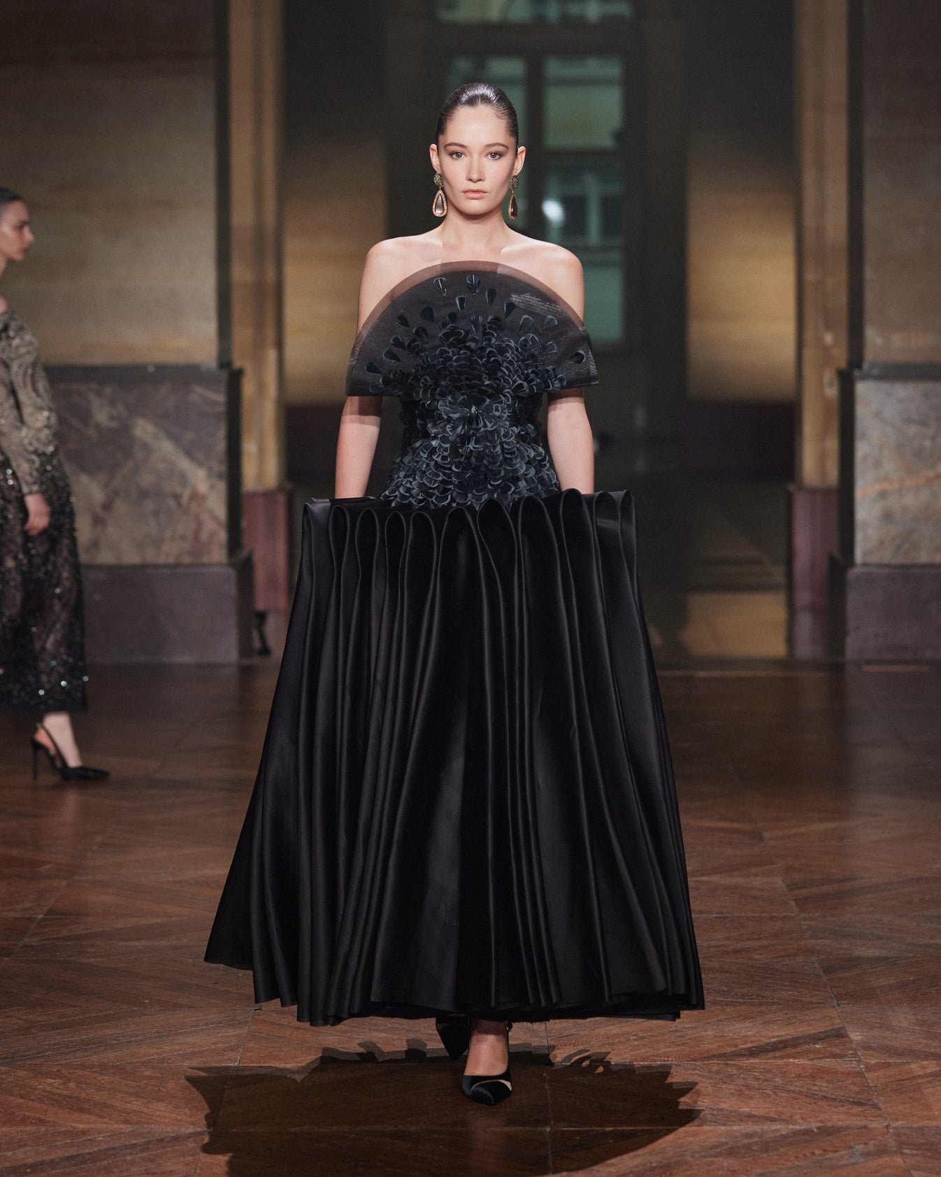 Modelo em vestido preto elegante com detalhes elaborados no busto, desfilando. Destaque para tendências da semana de alta-costura.