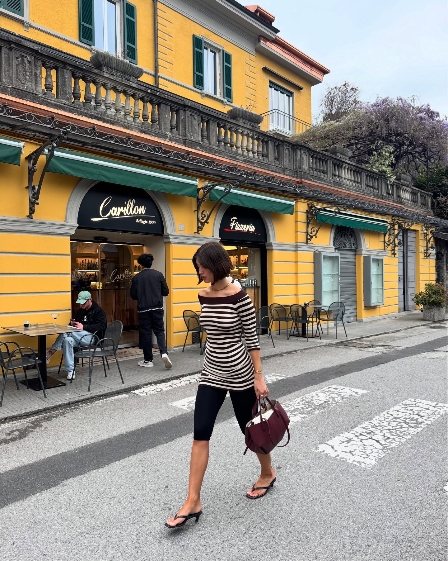 Mulher usa calça justa preta e blusa listrada caminhando em frente a prédio amarelo vibrante.