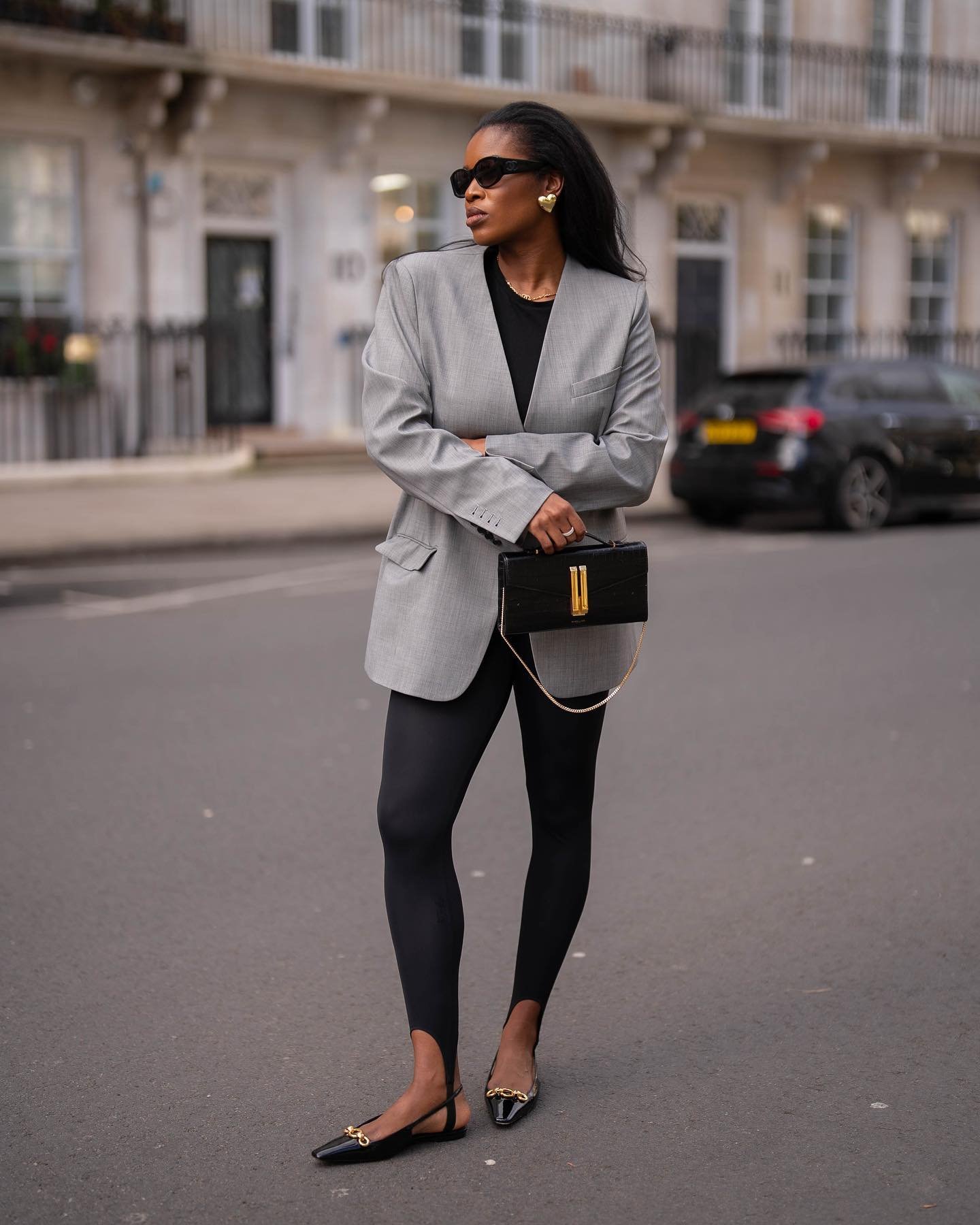 Mulher com blazer cinza e legging preta em rua urbana, segurando bolsa preta. Representa a ideia de tendências calça legging.
