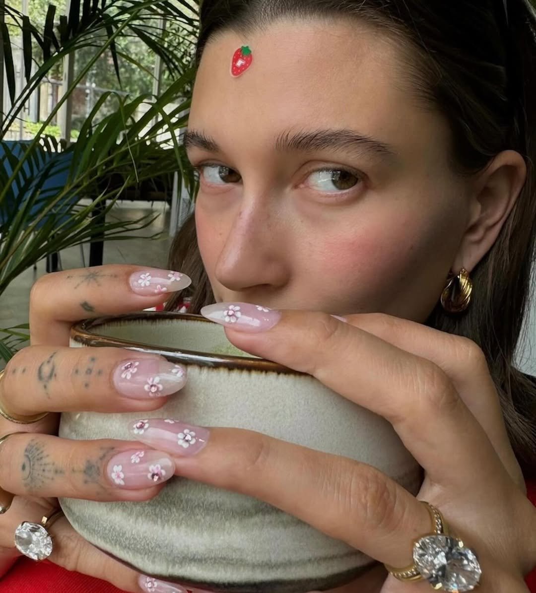 Unhas decoradas com pequenas flores brancas e rosas, segurando uma tigela enquanto uma pessoa olha para a câmera.