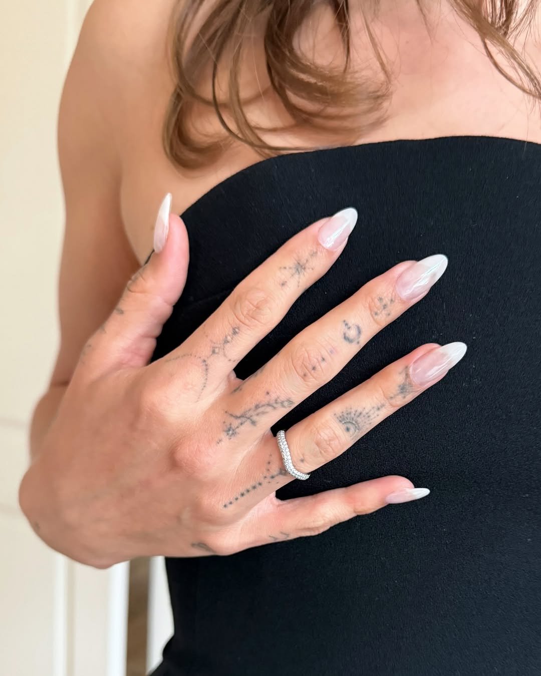 Unhas compridas e claras com anéis prateados e tatuagens delicadas nas mãos sobre tecido preto.