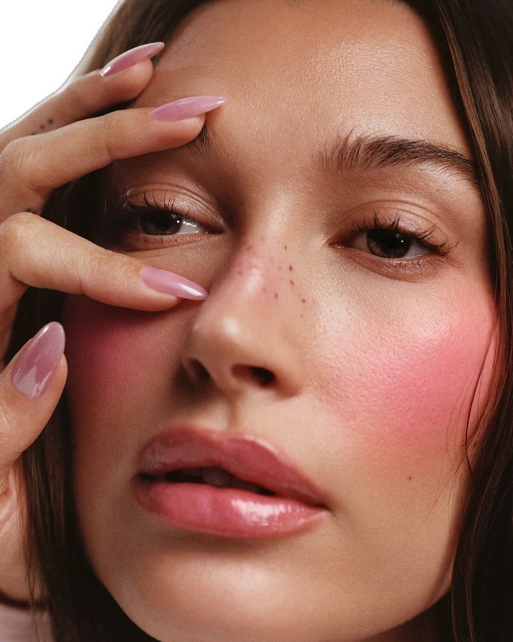 Unhas rosadas e brilhantes destacam-se em mão tocando suavemente o rosto feminino.