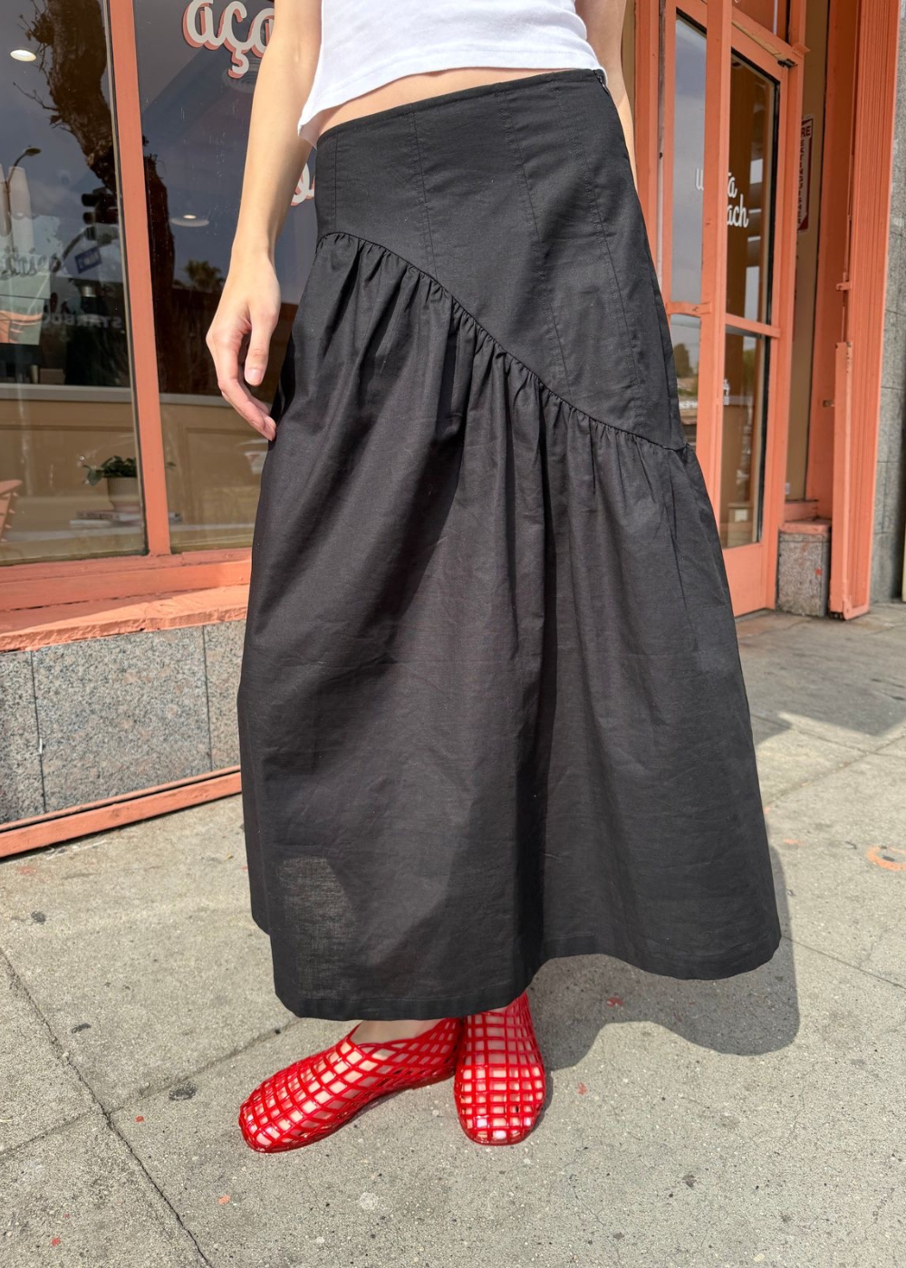 Saia preta estilo drop waist com sapatos vermelhos vazados, em frente a uma vitrine.