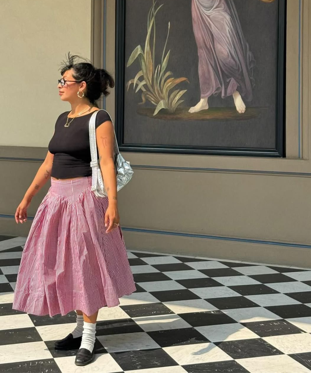 Pessoa com saia xadrez rosa de cintura rebaixada, camiseta preta e bolsa prata, em piso quadriculado. Tendência drop waist.