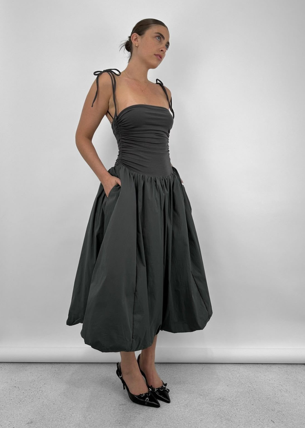 Vestido cinza escuro com cintura baixa, alças de amarrar e saia volumosa. Modelo usa sapatos pretos elegantes.