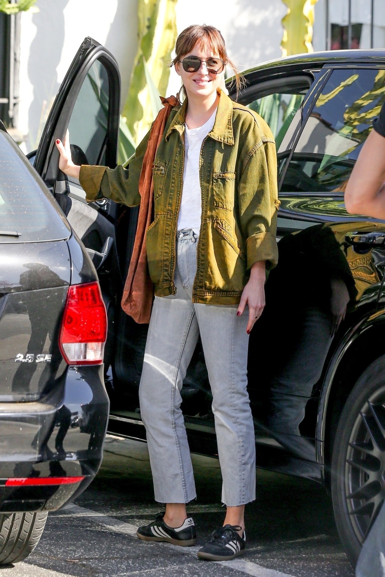 Pessoa usando jaqueta verde e calça jeans, saindo de um carro. Ela veste tênis estilosos pretos.