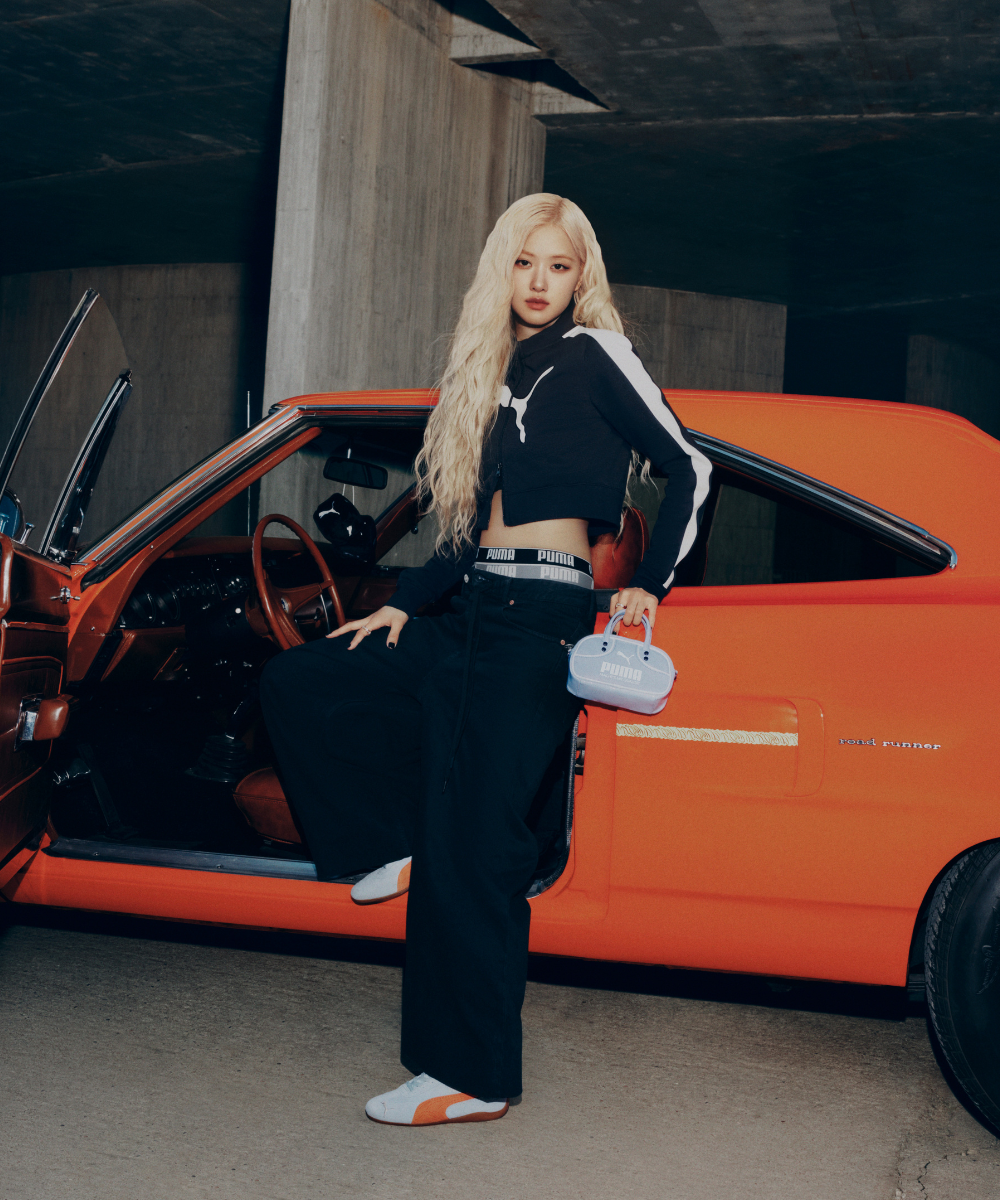 Mulher ao lado de um carro laranja, usando roupa esportiva Puma e segurando bolsa. Sneakers Speedcat nos pés.