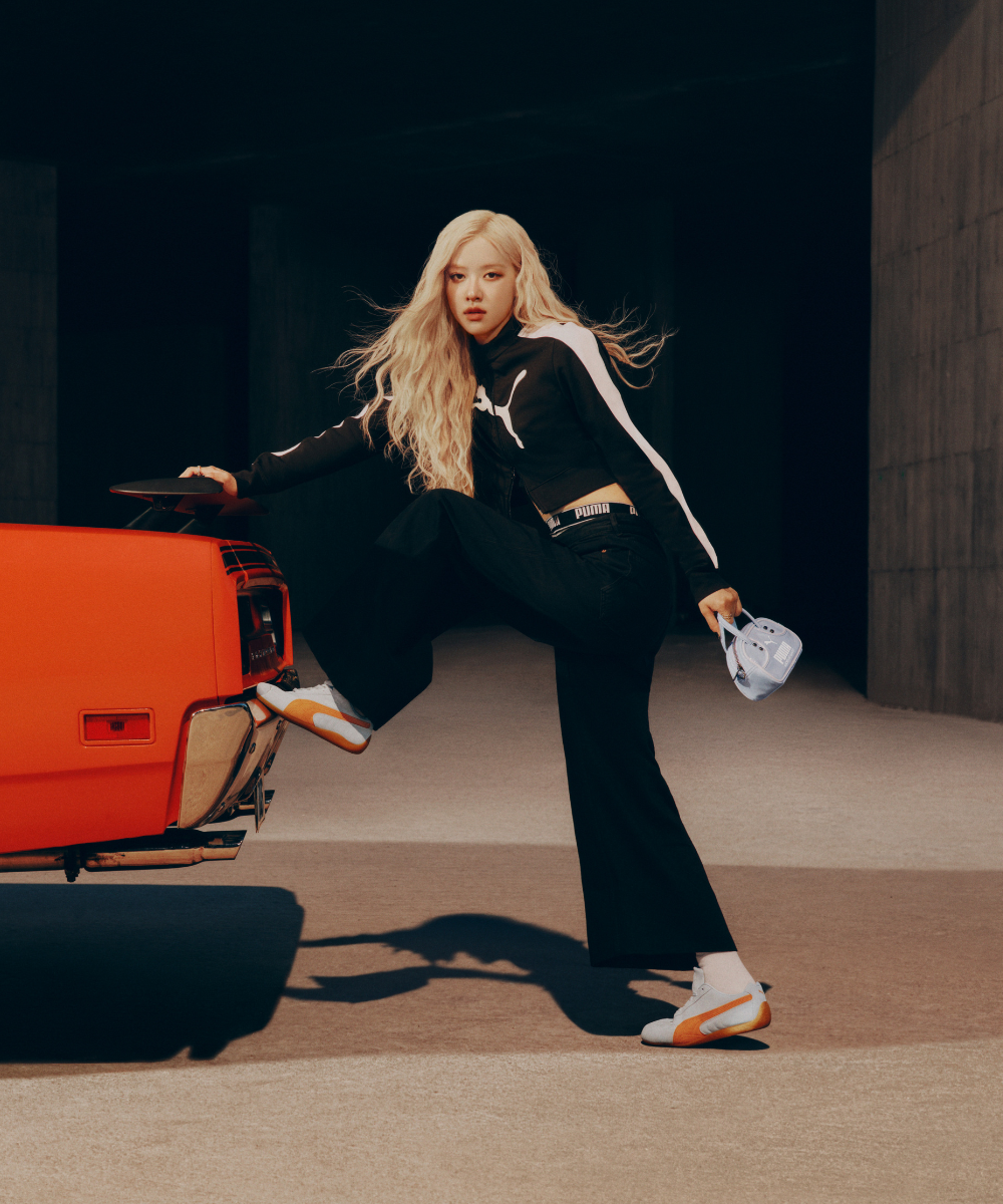 Mulher loira com roupa esportiva preta e tênis Speedcat, ao lado de um carro laranja. Ela segura um boné azul.