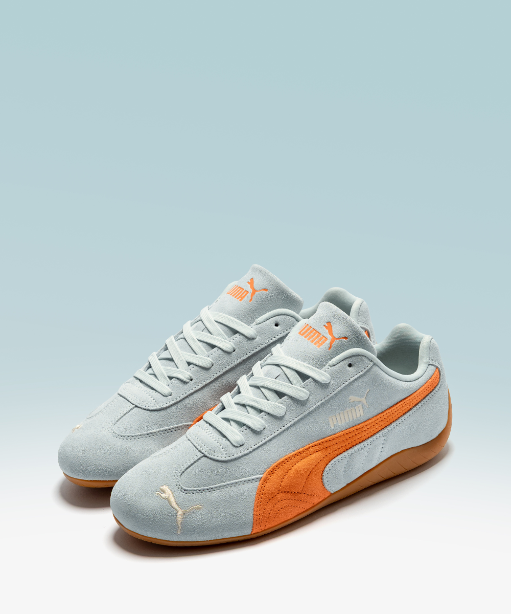 Tênis esportivos azul claro com detalhes laranja da linha Speedcat em fundo azul suave.