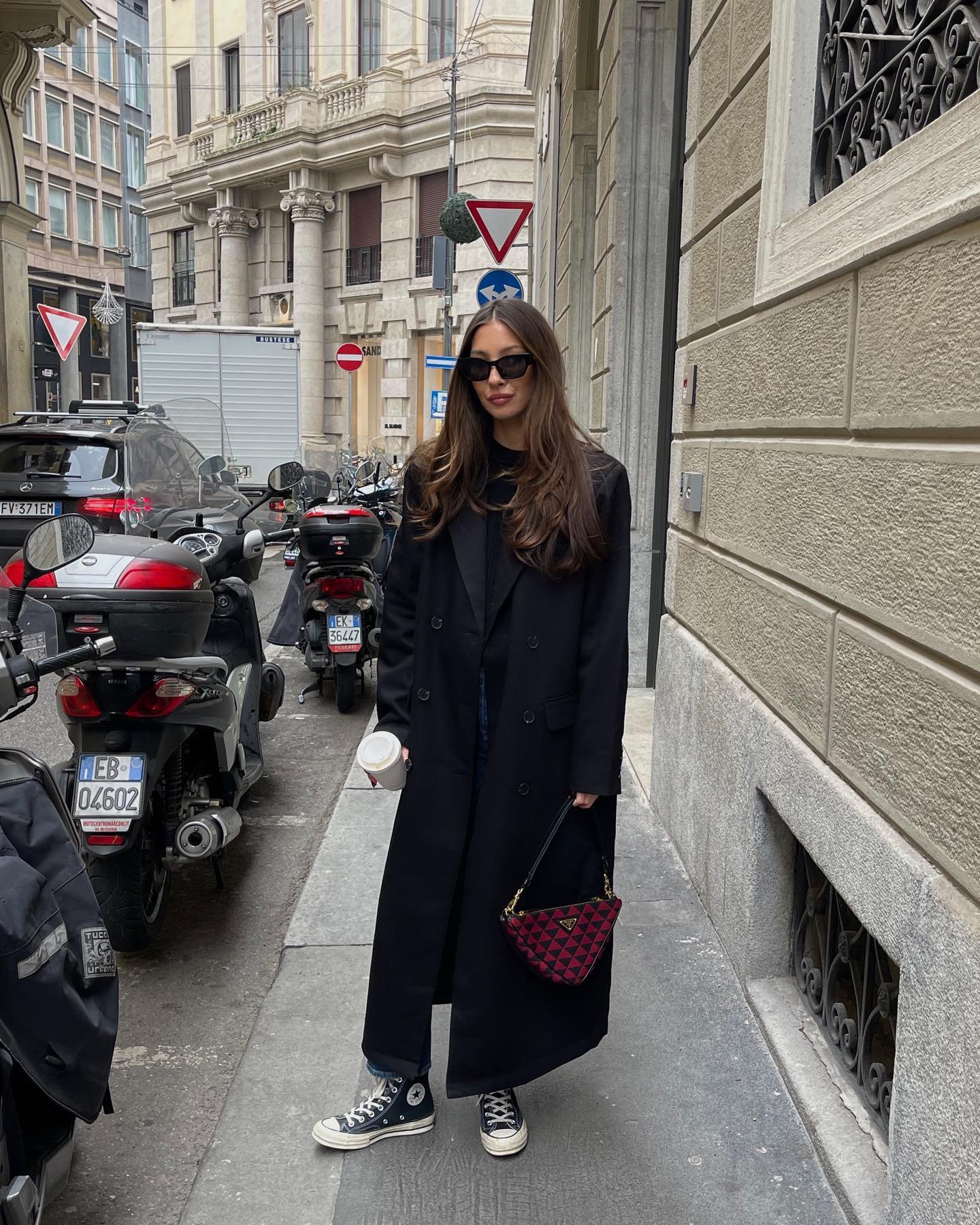 Mulher com casaco preto, óculos escuros e tênis estilo clássico, segura café em uma rua urbana. Tênis em tendência para 2025.