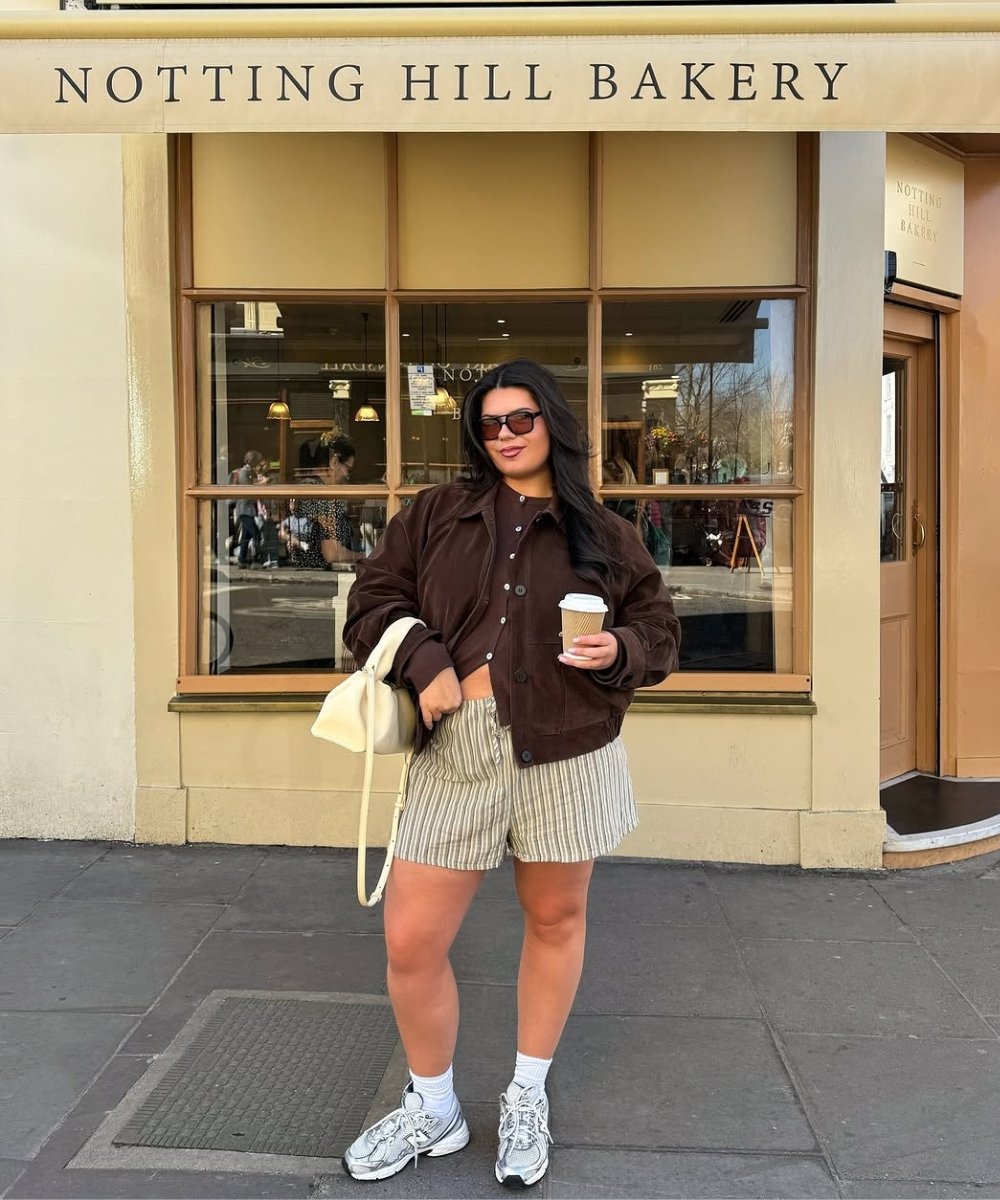 Jovem com shorts listrados e jaqueta marrom, usando tênis modernos. Segura café em frente a padaria.