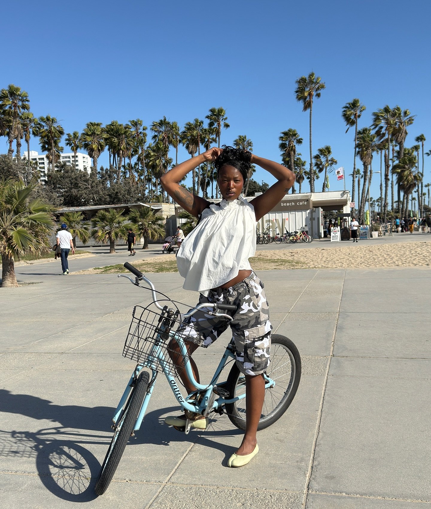 Pessoa de pé ao lado de uma bicicleta em local com palmeiras e céu azul. A 