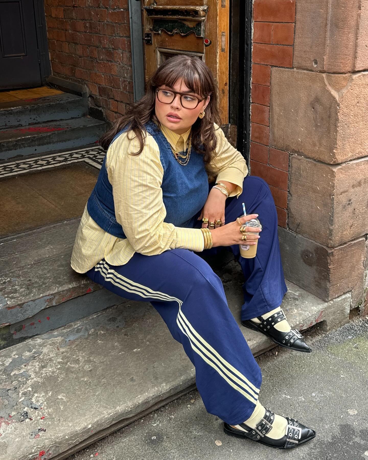 Uma pessoa sentada em degrau, vestindo camisa amarela, colete azul e calças com listras, segura bebida. O sapato inclui detalhes que exemplificam uma escolha não usual, reforçando a teoria do sapato errado como destaque.