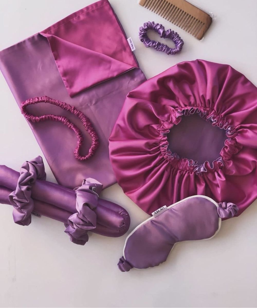 Conjunto de acessórios de cetim rosa e roxo, incluindo touca de cetim, fronha, máscara de dormir, elásticos e pente de madeira.