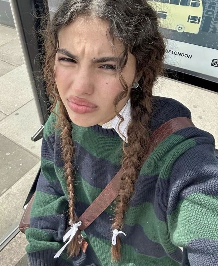 Jovem com tranças soltas e efeito desalinhado. Ela está usando um suéter listrado, sentada em um ponto de ônibus. Penteados sem tração