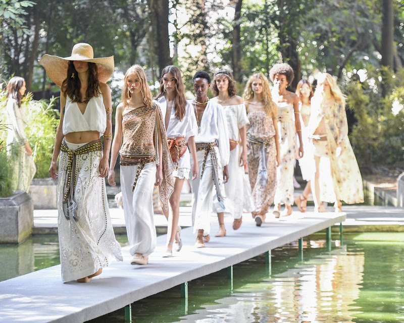 Desfile de moda ao ar livre com modelos usando roupas leves e acessórios boho, refletindo o estilo de Maria Filó.