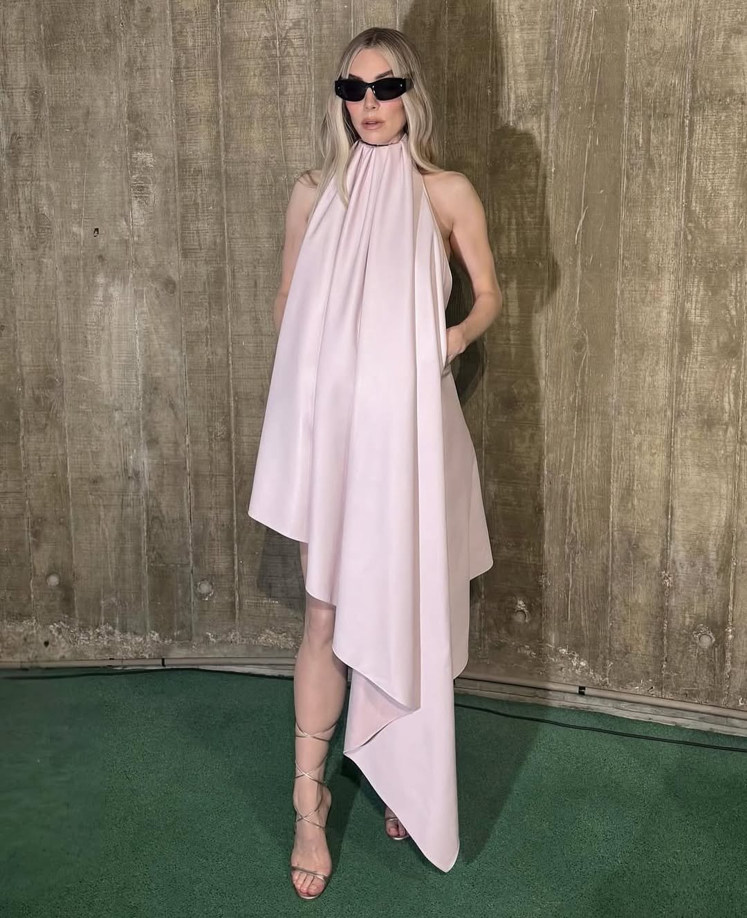 Vanessa Kirby de vestido rosa e óculos escuros posa em frente a parede de madeira.