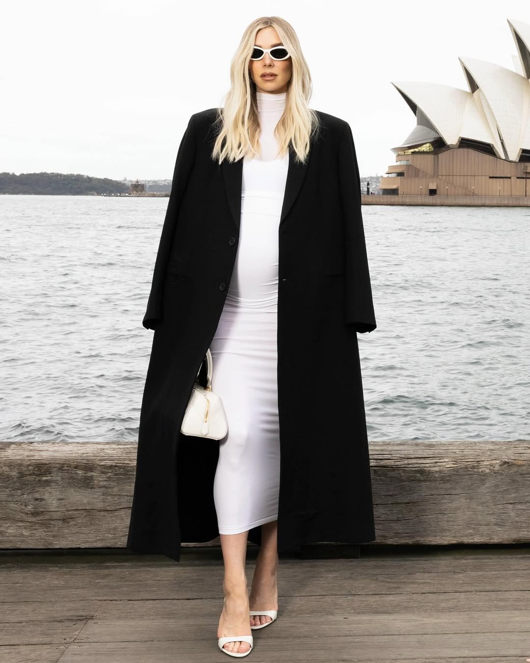 Vanessa Kirby com vestido branco e casaco preto, próximo à água e um edifício icônico ao fundo.