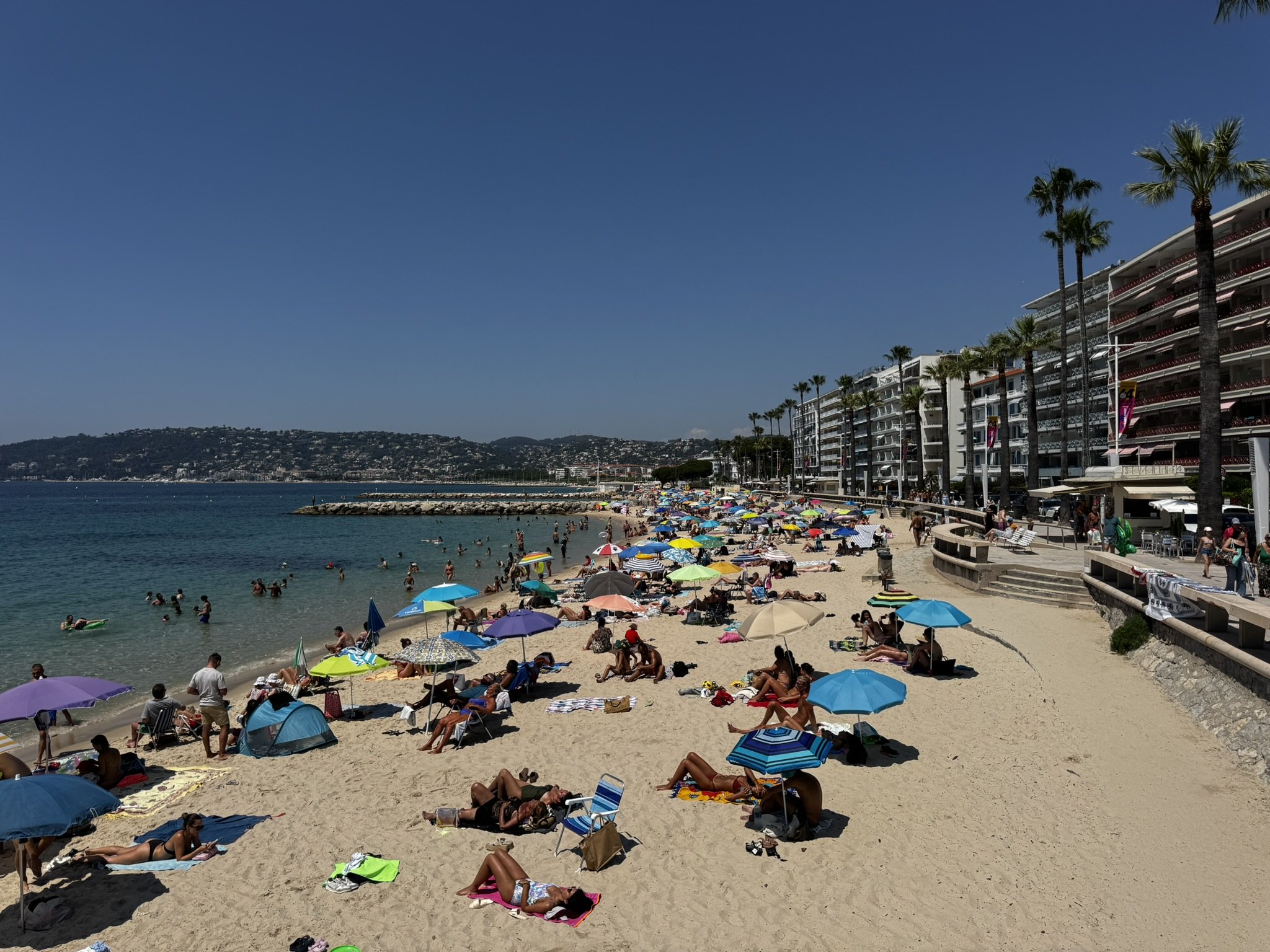 Praia movimentada com pessoas sob guarda-sóis coloridos, litoral ensolarado e prédios ao fundo, típico do verão europeu.