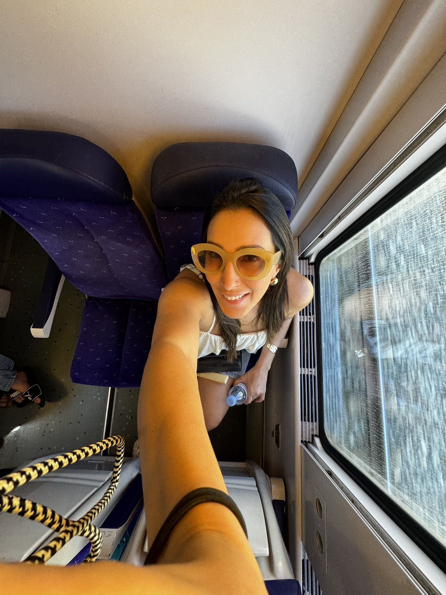 Um passageiro sorri em um trem, aproveitando o verão europeu com óculos de sol e roupa leve.