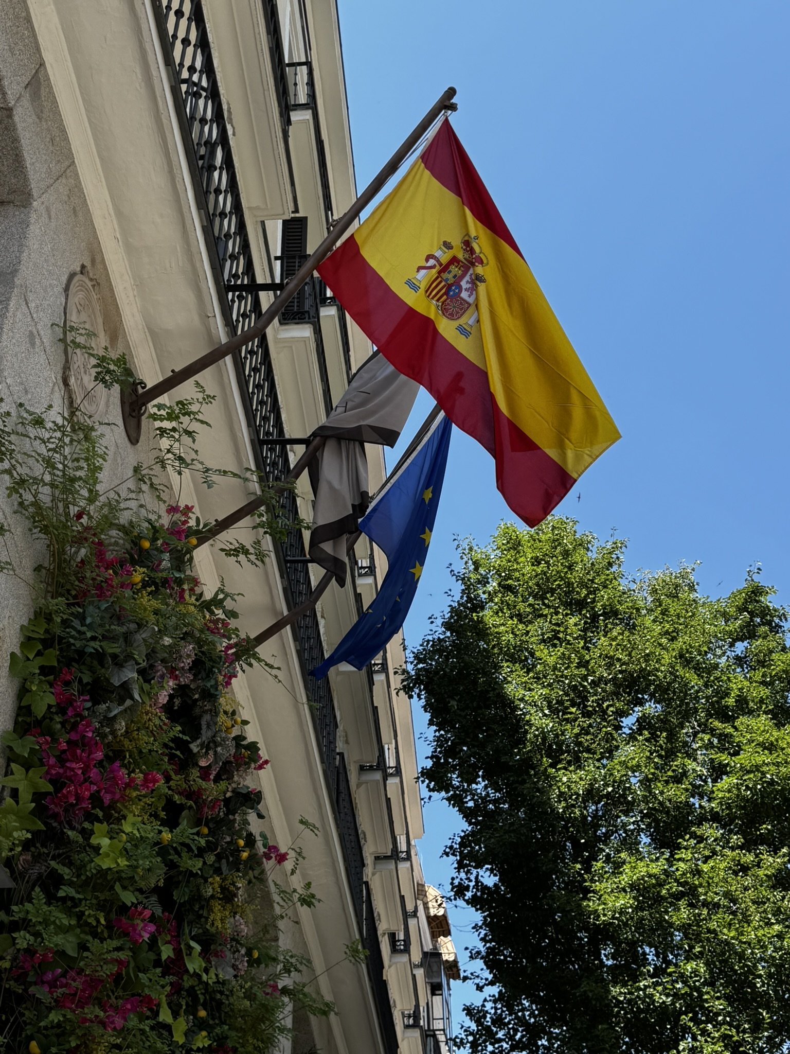 Bandeira da Espanha ao lado da bandeira da UE em um prédio, céu azul sugere verão europeu.