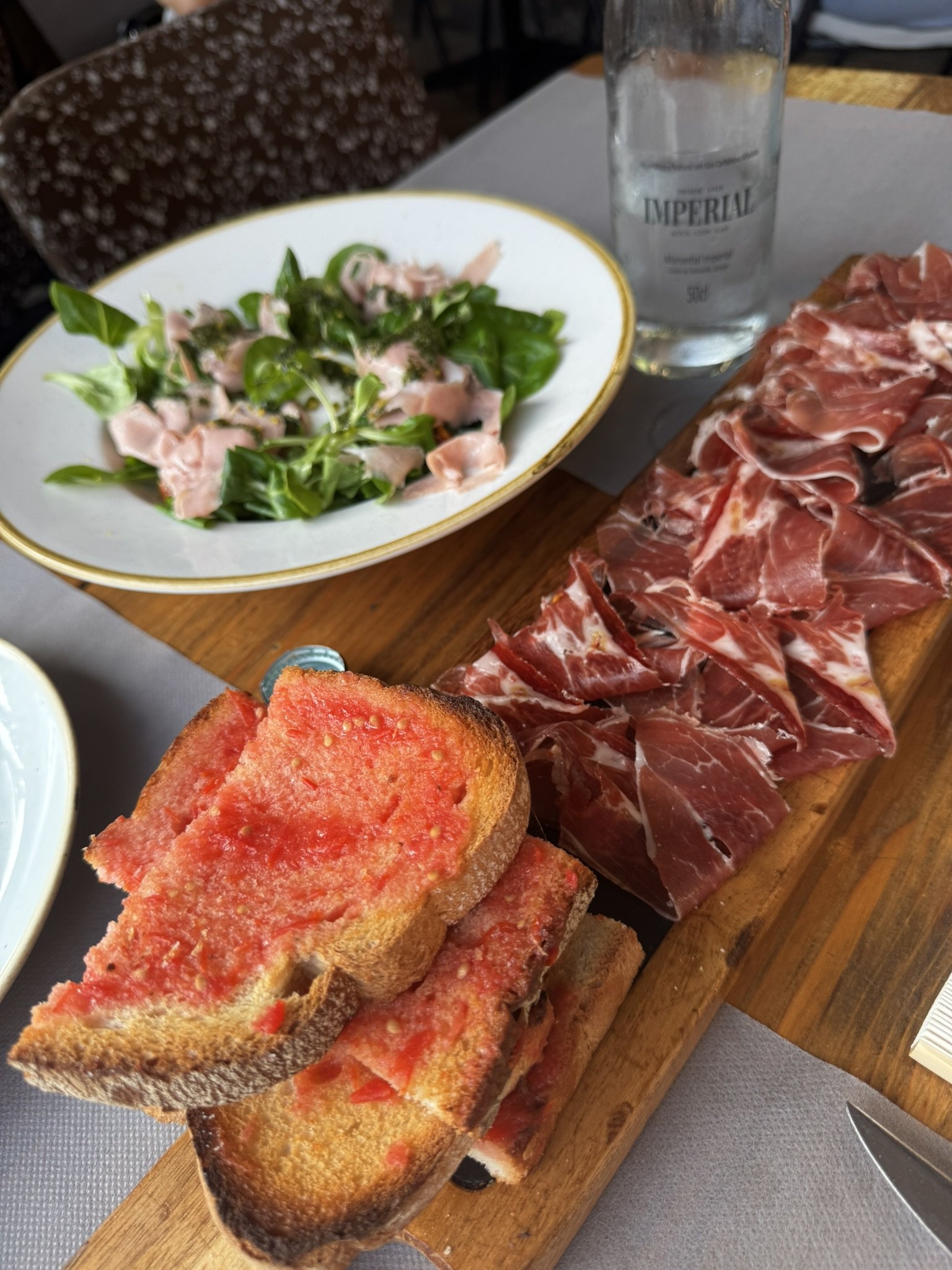 Mesa com presunto, pão com tomate e salada, evocando um almoço leve típico do verão europeu.