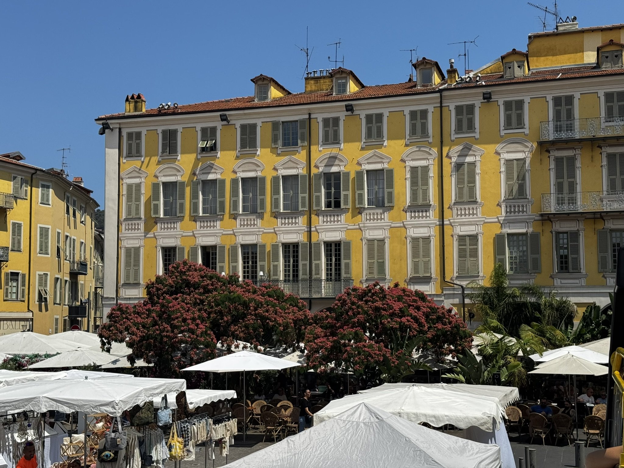 Feiras ao ar livre com árvores floridas em frente a prédios amarelos típicos do verão europeu.