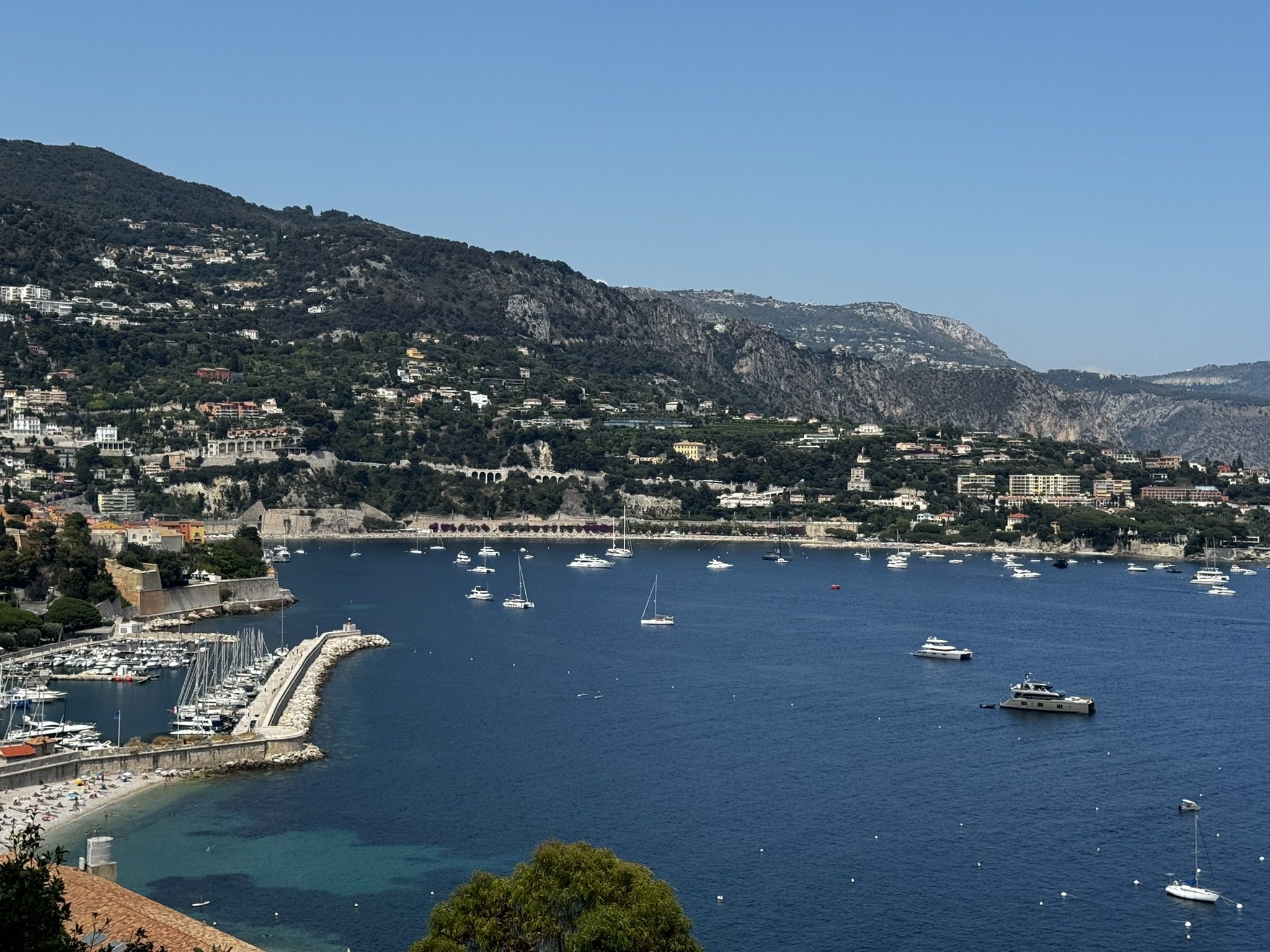 Costa europeia com montanhas, cidade e marina sob céu azul, típica do verão europeu.