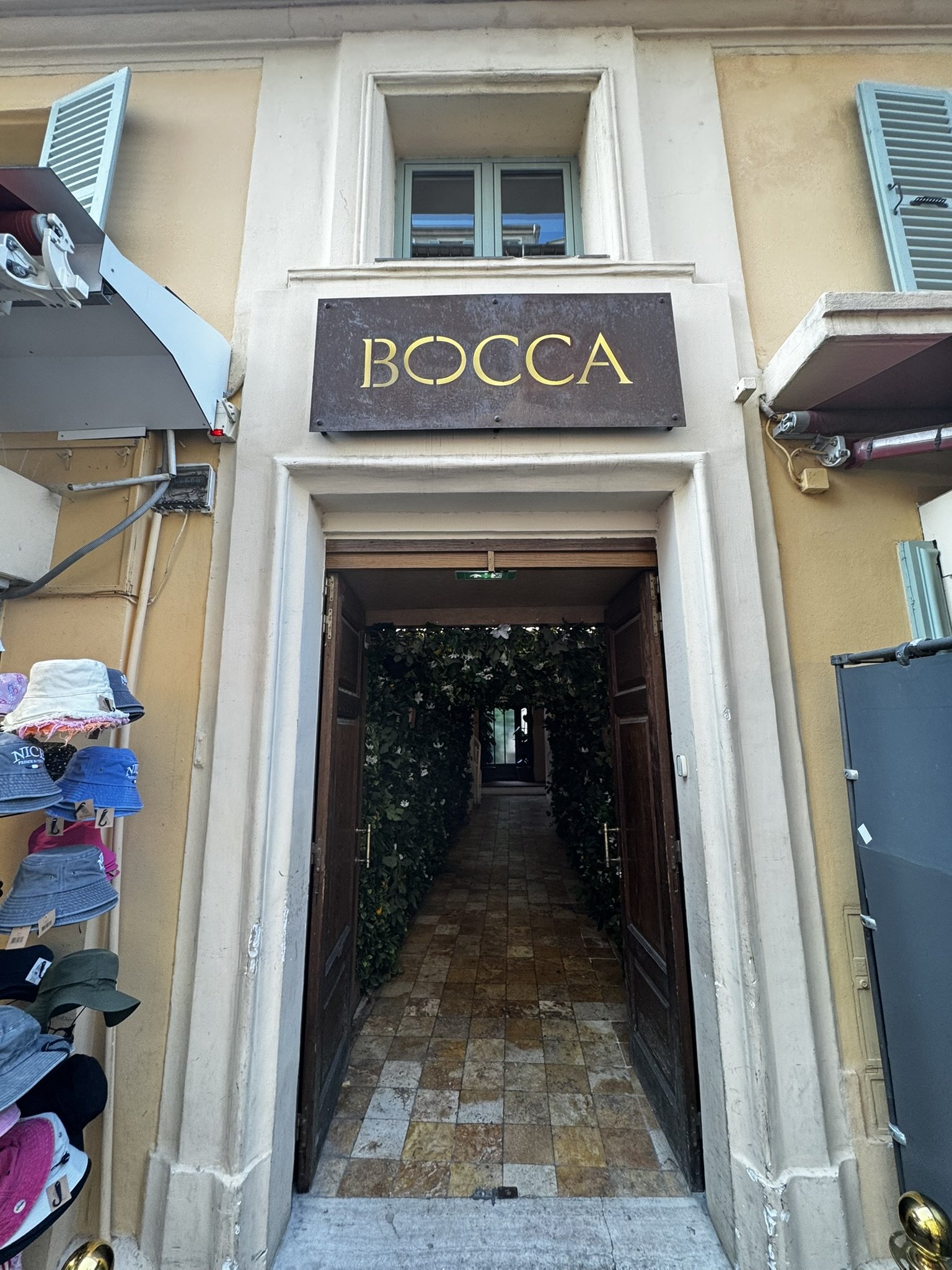 Entrada de loja chamada Bocca com paredes de plantas, evocando o charme do verão europeu.