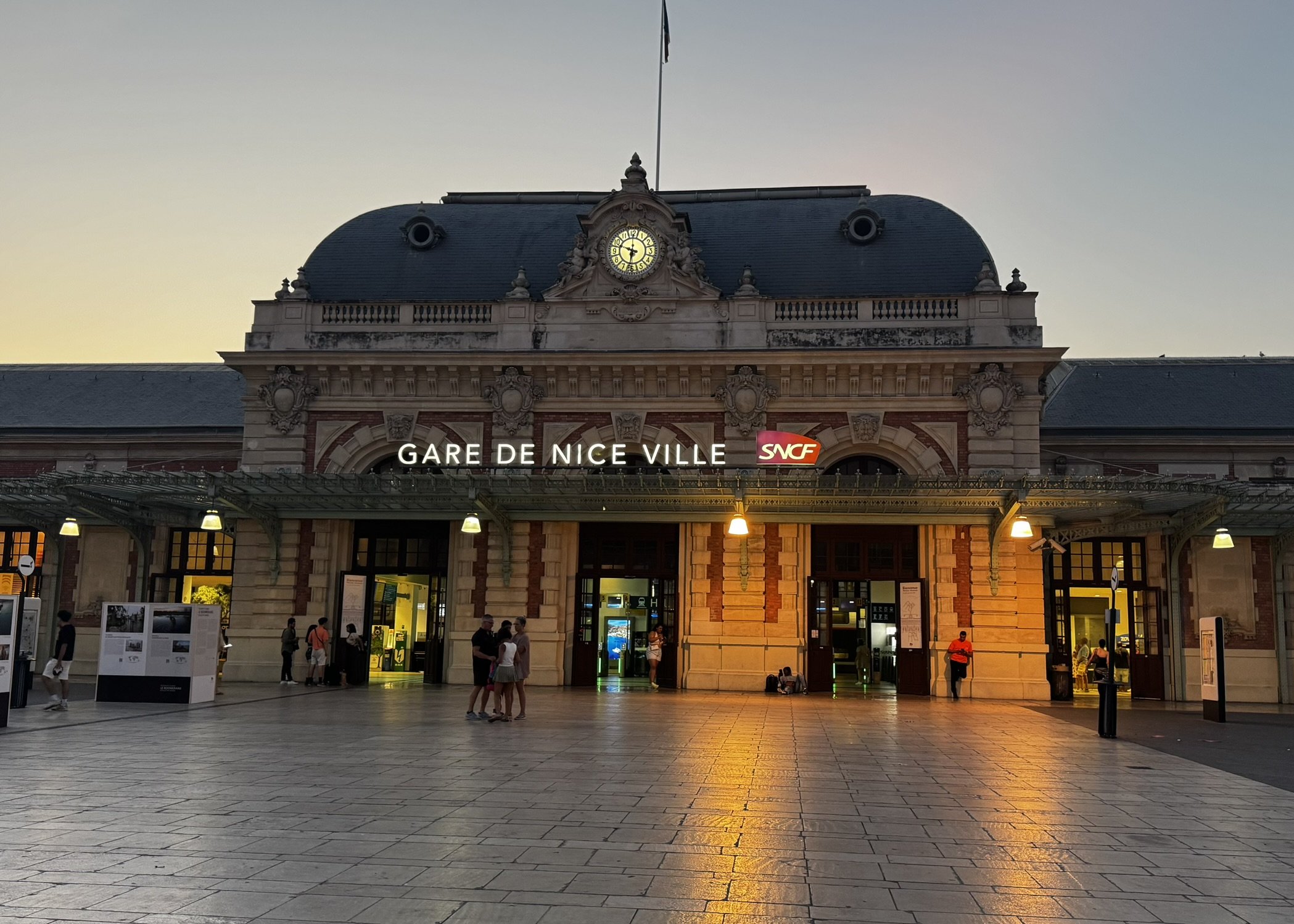 Estação de trem Gare de Nice-Ville ao entardecer, durante o verão europeu, com pessoas na praça.