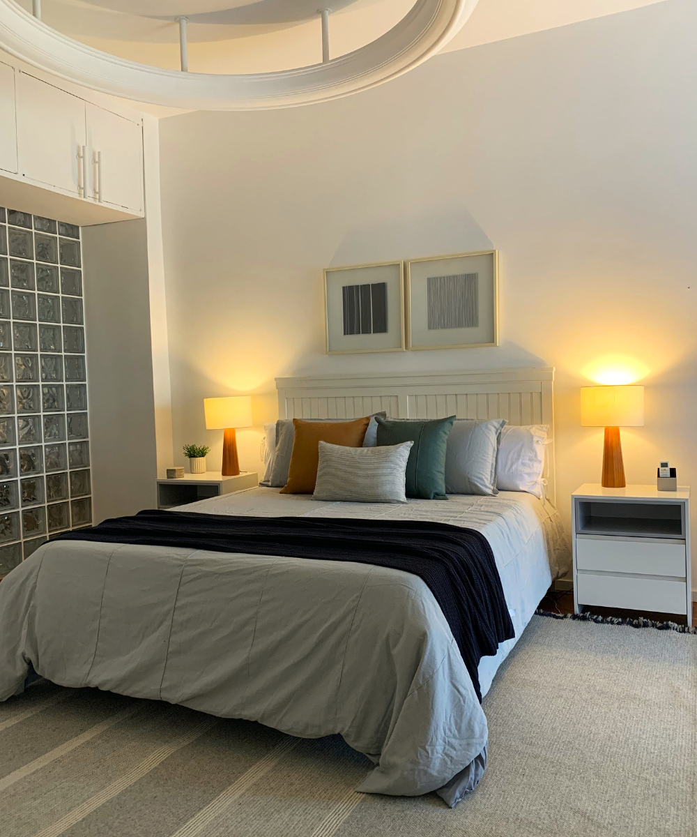 Quarto elegante com cama decorada, iluminação suave e quadros na parede, promovendo bem-estar e arquitetura acolhedora.
