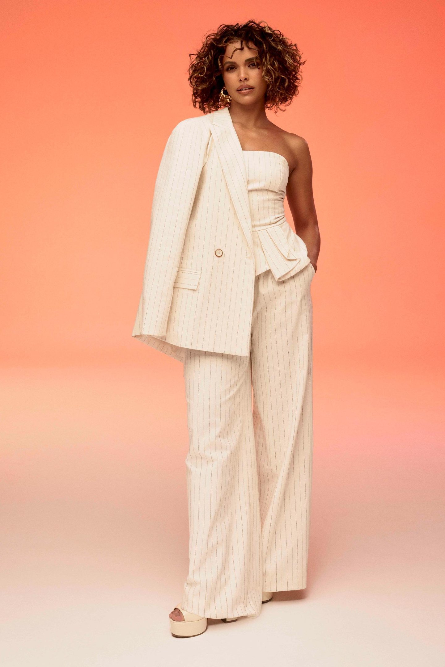 Modelo com terno bege listrado e cabelo cacheado, em fundo laranja suave. Estilo elegante e moderno. Flor de Lis.