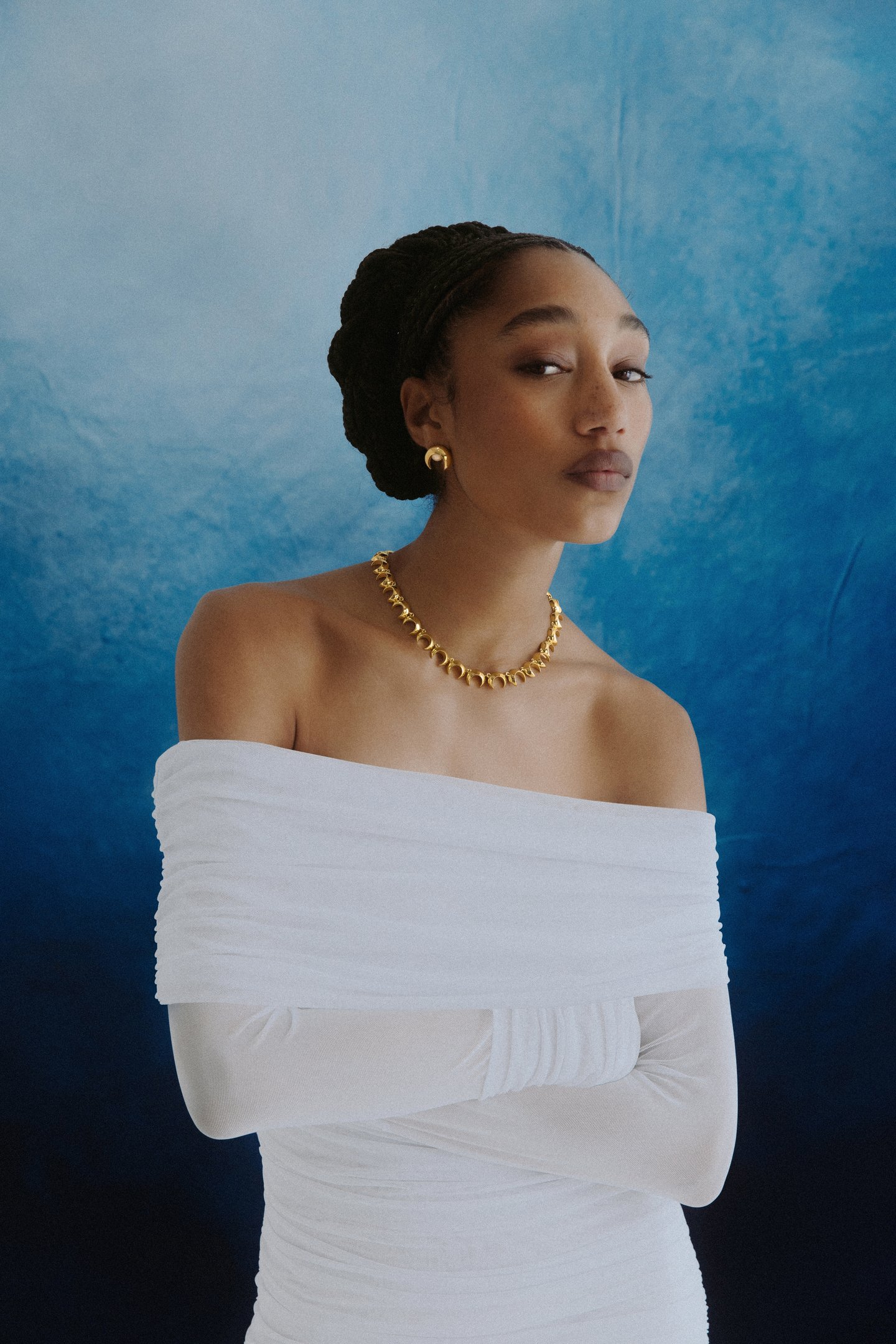 Mulher em vestido branco ombro a ombro, colar e brincos dourados, em fundo azul.