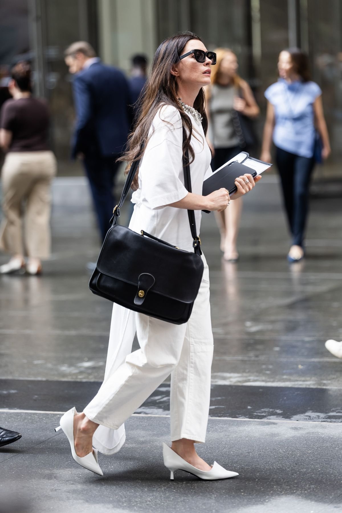 Vejo uma mulher caminhando na rua, vestindo roupa branca e carregando bolsa preta.