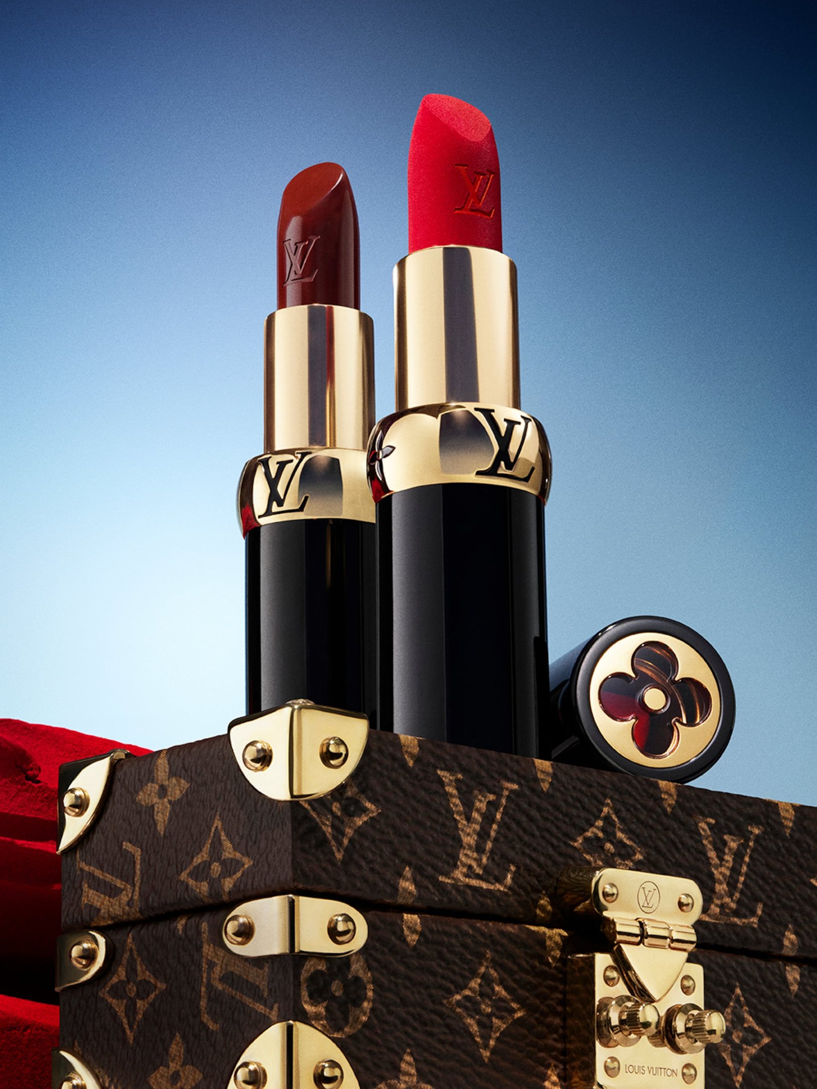 Dois batons Louis Vuitton em tons vermelho e marrom sobre caixa com detalhes dourados e logotipo da marca.